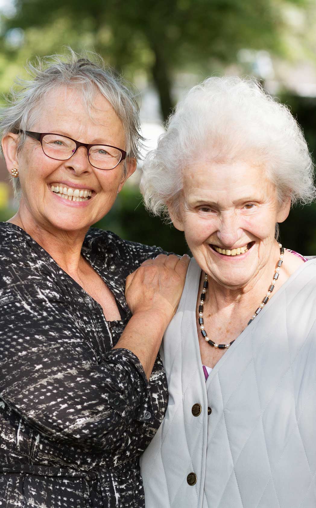 Two happy elderly women, one with her hand on the other's shoulder, smile for their photo to be taken.