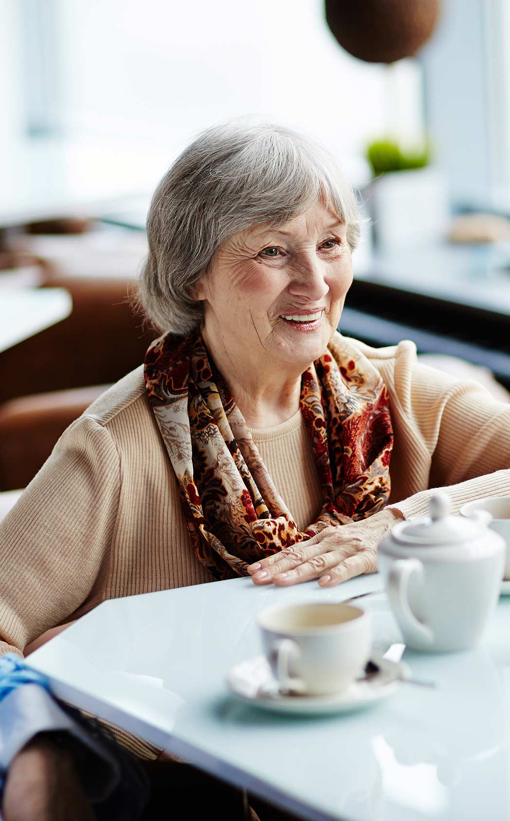 A senior lady smiles while sitting and speaking with someone over coffee.