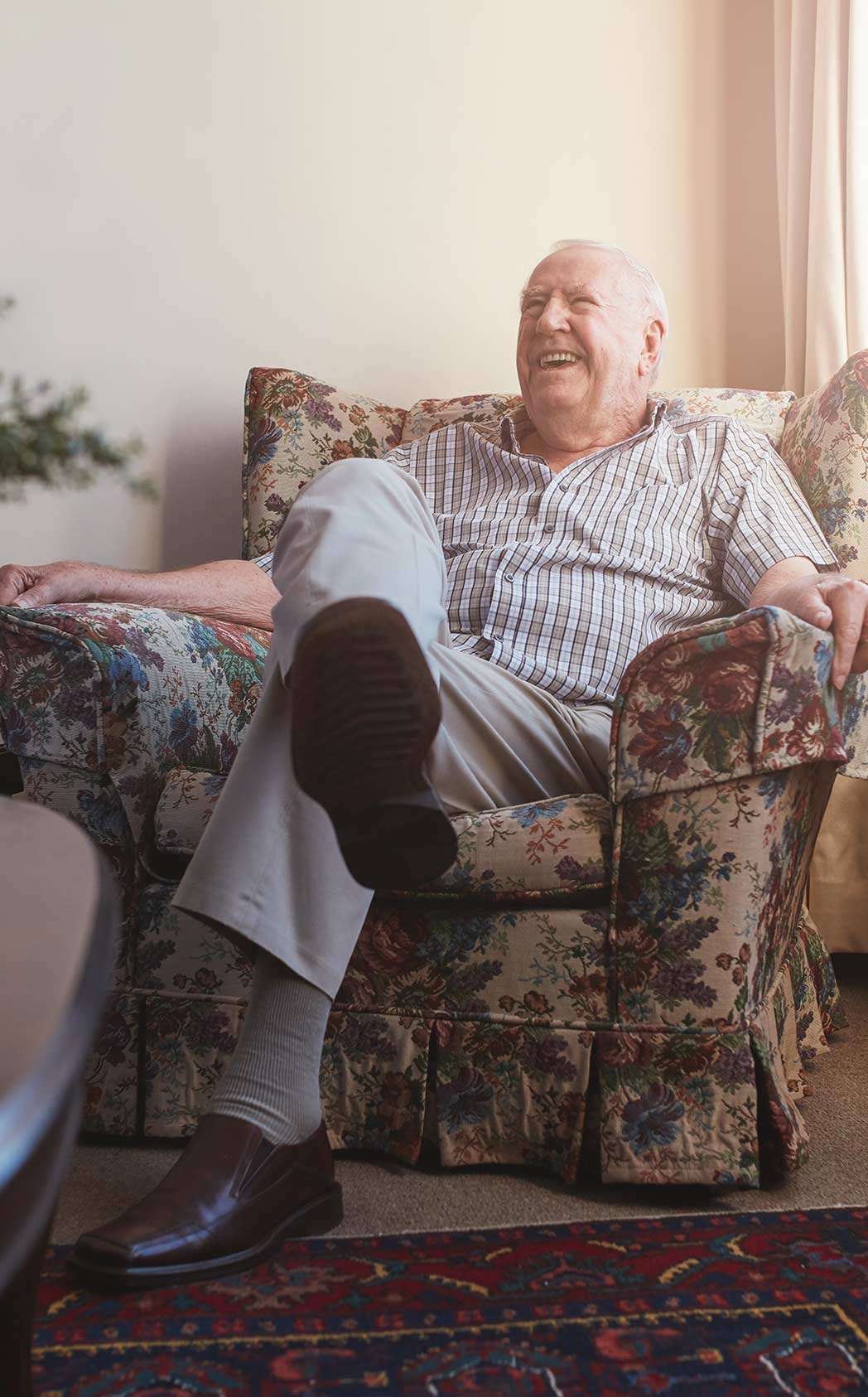 A view of a senior man sitting in a living room armchair with his legs crossed, laughing with the person visiting him.