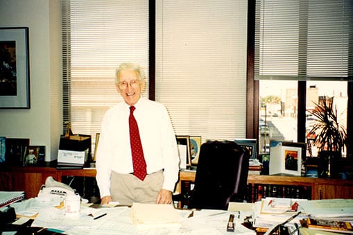 Bill R. Foster, Founder of Foster Hospitality Group stands behind his desk smiling for a photo