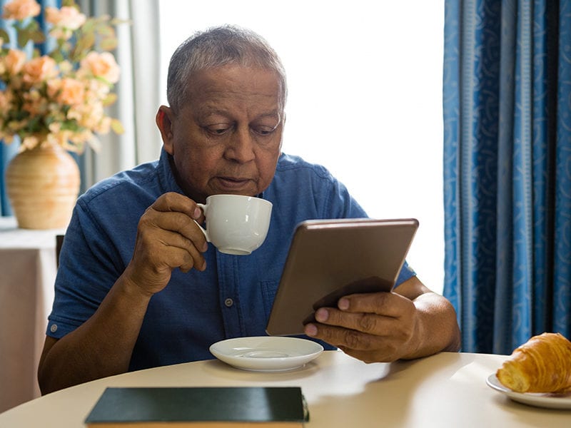 A man sits at a table and sips his coffee while checking his iPad.