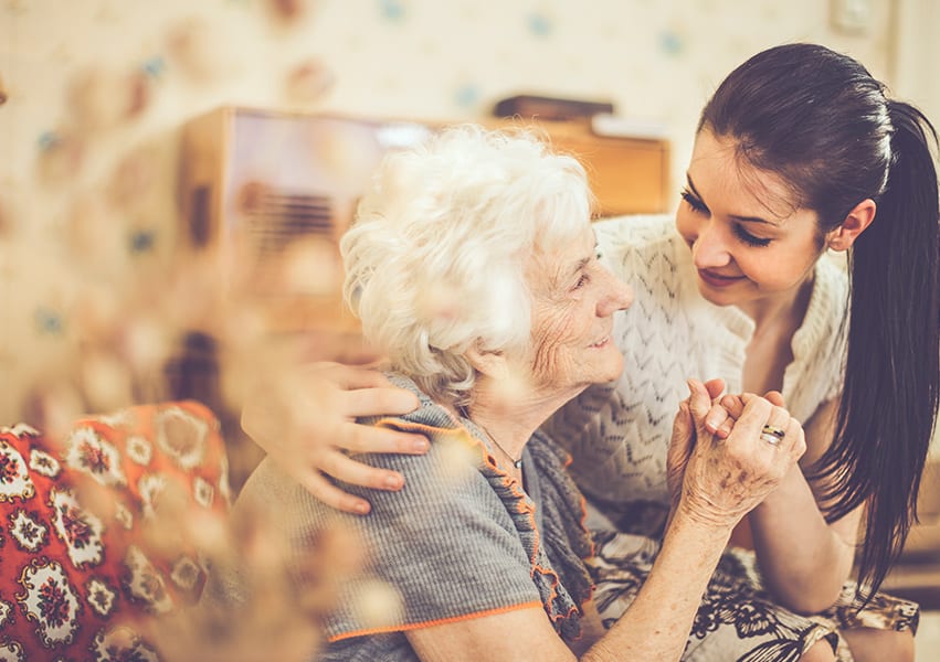 A grown or mostly grown grandchild sits on the lap of her grandmother, looking into each others eyes and clasping hands.