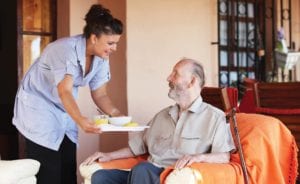 A Culpepper Place employee brings nutritious food and drink on a tray to a resident who is sitting in an armchair.