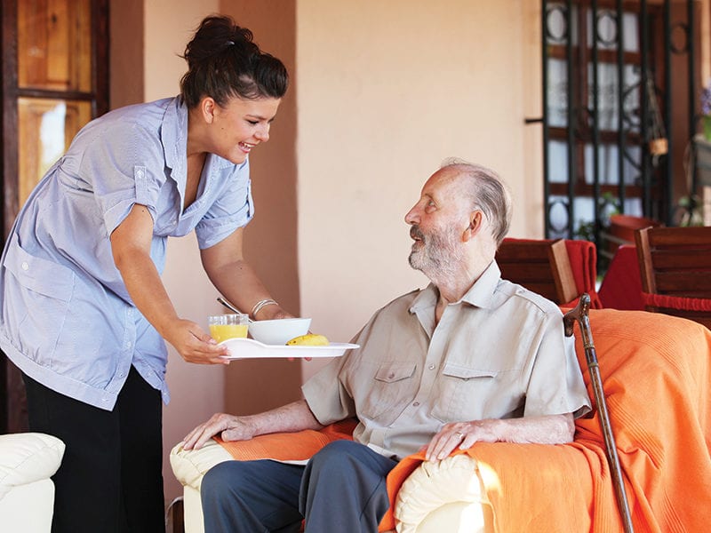 A Culpepper Place employee brings nutritious food and drink on a tray to a resident who is sitting in an armchair.