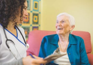 A clinician wearing scrubs and stethoscope holds a clipboard and speaks with a happy woman resident.