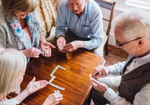 Four assisted living residents are playing dominos together in a recreation room where board games are available.