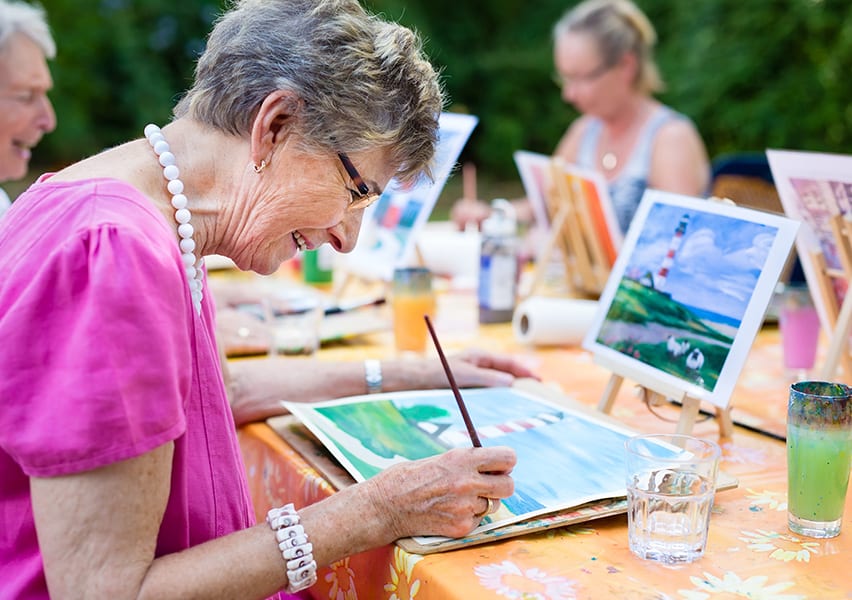 A woman resident paints a lighthouse from an example photo while doing this activity outdoors with others.