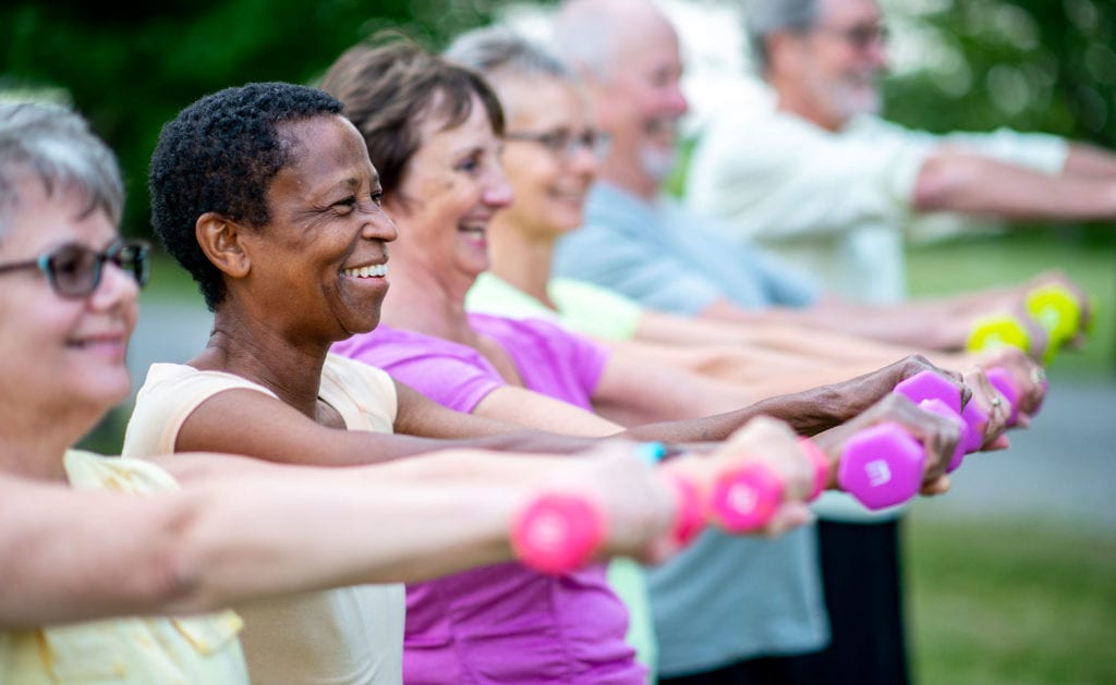 A smiling group of individuals stand exercising outdoors with free weights extended in front of them.
