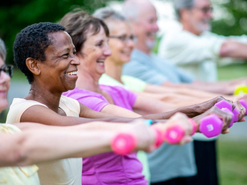 A smiling group of individuals stand exercising outdoors with free weights extended in front of them.