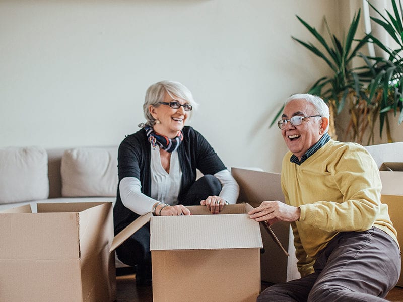 Two seniors moving into their private assisted living apartment unpack and have a good time as they smile from the floor.