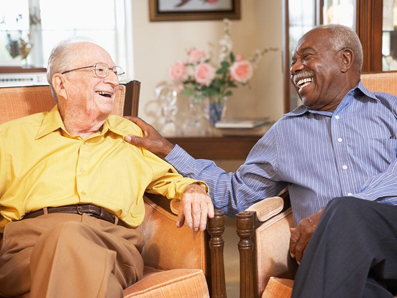 Two elderly male residents who are laughing and smiling are having a good time being social with one another as they sit in a living area.