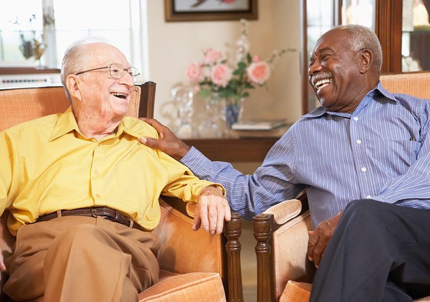 Two elderly male residents who are laughing and smiling are having a good time being social with one another as they sit in a living area.